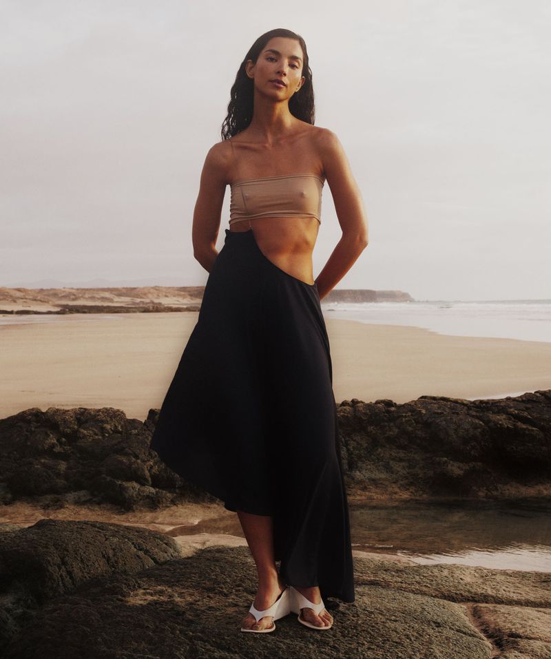 Woman in a bandeau top and skirt stands on a rocky beach near the ocean on an overcast day in a fashion style photograph.