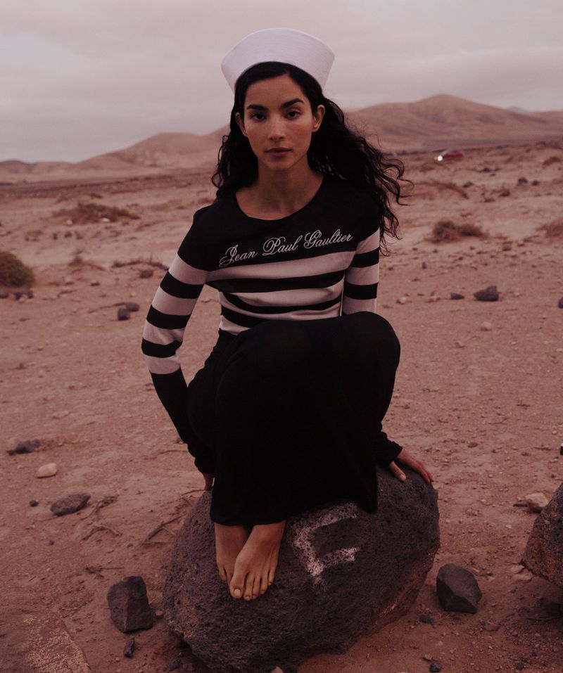 Woman wearing a sailor hat and striped shirt, sitting on a rock in the desert landscape, looking at the camera. Moody outdoor lighting.