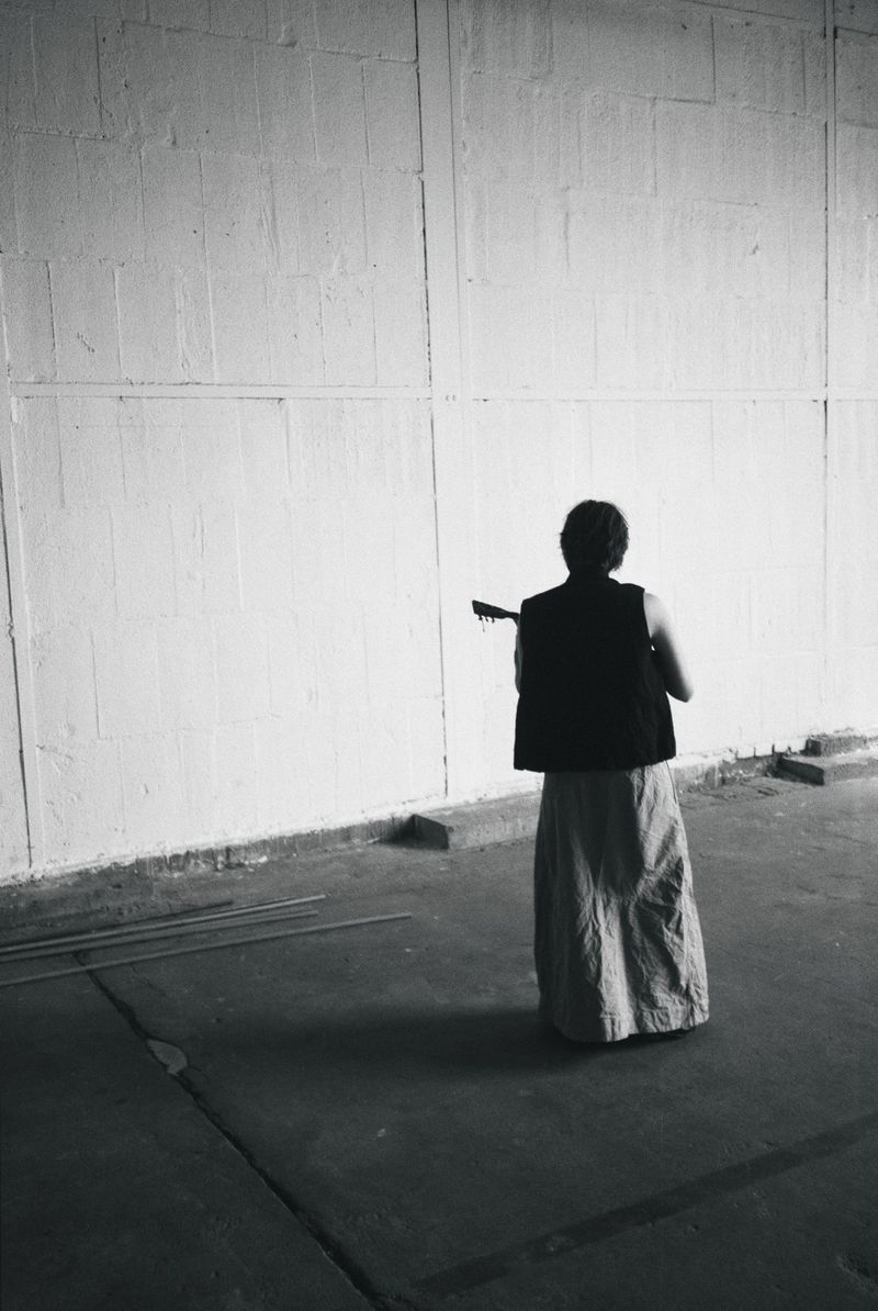 Rear view of a figure in a black vest and long textured skirt holding a stringed instrument in a spacious concrete warehouse.