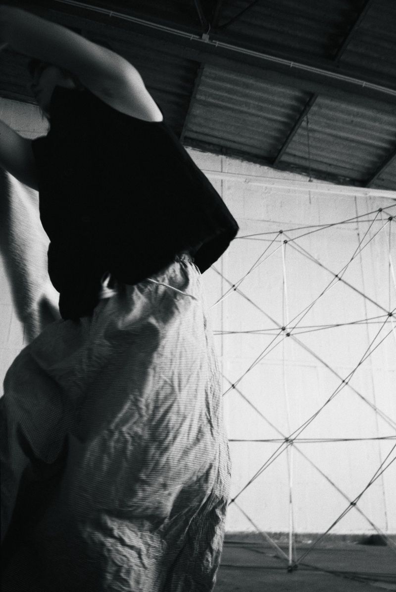 Low-angle black and white shot of a person in textured baggy trousers and a black top standing in an industrial space with a wire structure.