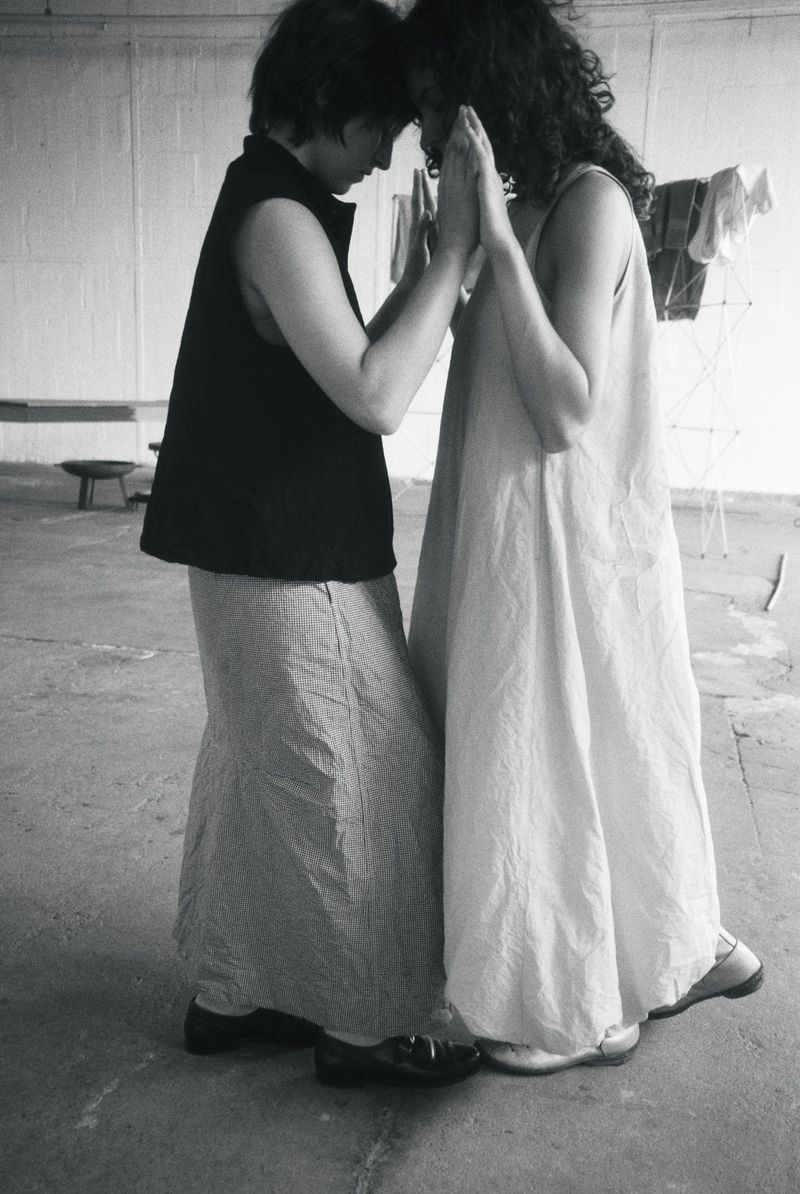 Two women standing face-to-face touching palms and foreheads in monochrome, wearing a sleeveless maxi dress and a dark vest.