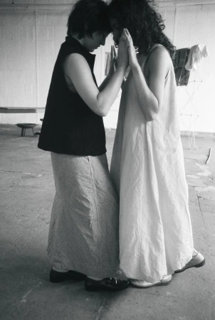 Two women standing face-to-face touching palms and foreheads in monochrome, wearing a sleeveless maxi dress and a dark vest.