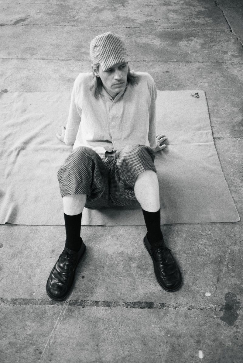 Model wearing a patterned cap, light shirt, patterned shorts, black socks, and shoes seated on a mat on concrete.
