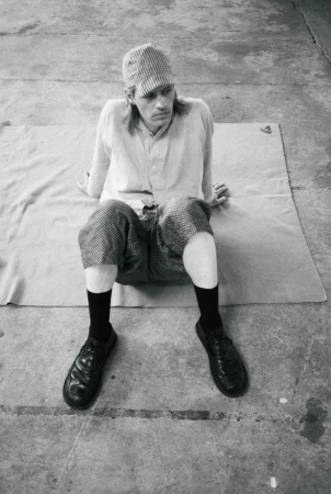 Model wearing a patterned cap, light shirt, patterned shorts, black socks, and shoes seated on a mat on concrete.