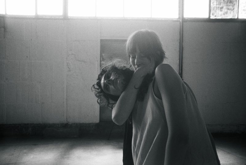 Black and white photo of two people closely embracing inside an industrial warehouse space with upper windows.