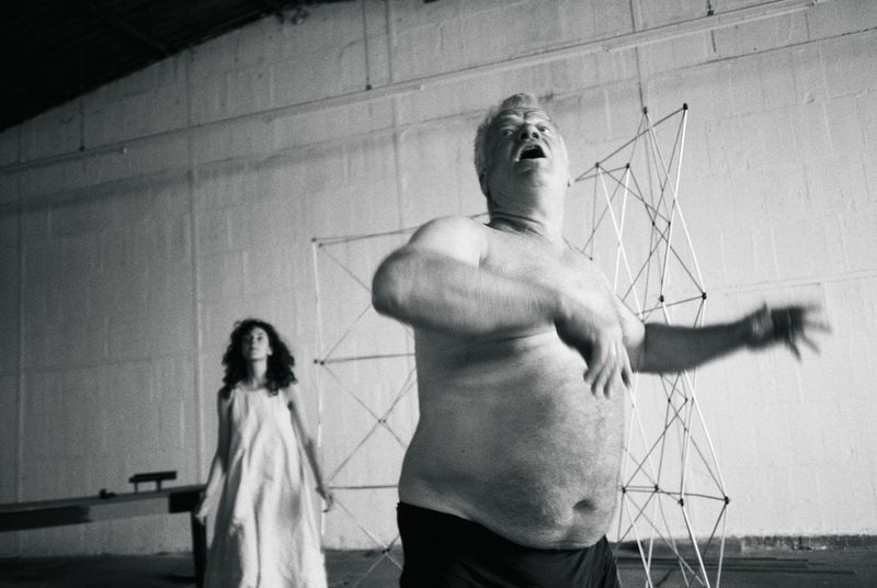 Black and white photo of a shirtless mature man gesturing dynamically in a warehouse studio next to a standing woman in a white dress.