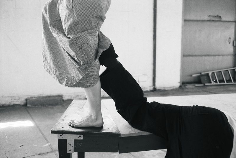 Black and white photo of a bare foot on a wooden bench beside a reclining figure in dark trousers within an industrial warehouse space.