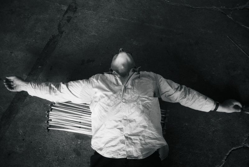 Monochrome overhead shot of a person lying on a concrete floor with arms outstretched, wearing a light wrinkled button-up shirt.