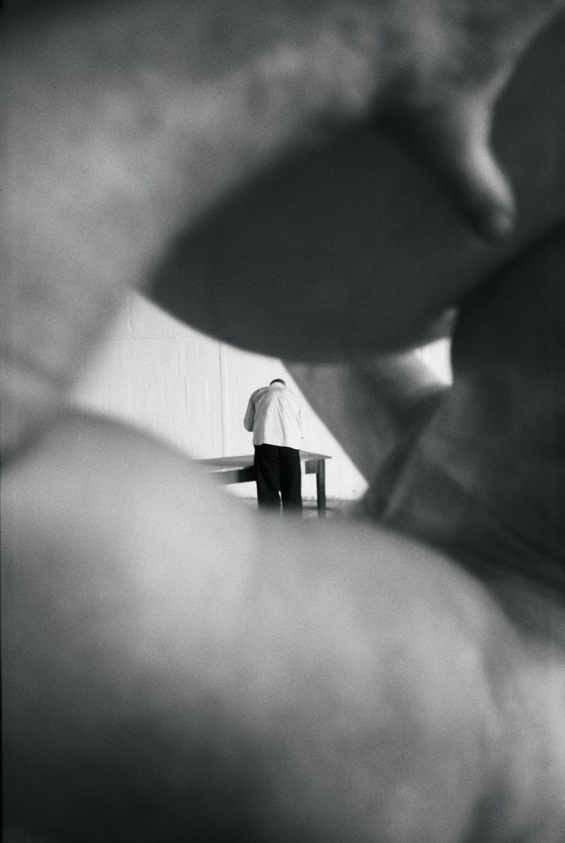 Monochrome shot of an adult male standing at a table near a white wall, framed by blurred human form. Interior scene.