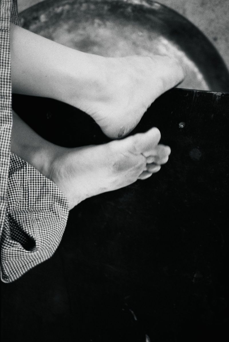 Close-up black and white photograph of bare feet resting over a dark surface beside fine-check patterned trouser fabric.
