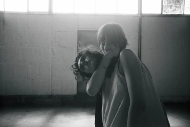 Black and white photo of two people closely embracing inside an industrial warehouse space with upper windows.