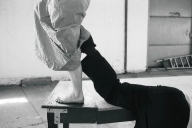 Black and white photo of a bare foot on a wooden bench beside a reclining figure in dark trousers within an industrial warehouse space.