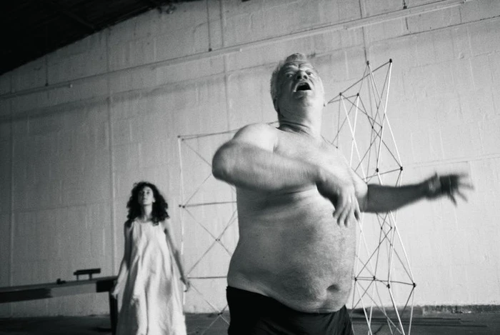 Black and white photo of a shirtless mature man gesturing dynamically in a warehouse studio next to a standing woman in a white dress.