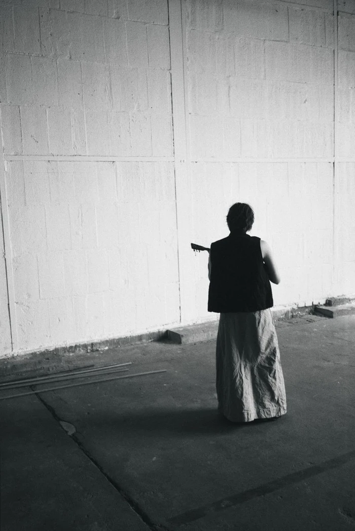 Rear view of a figure in a black vest and long textured skirt holding a stringed instrument in a spacious concrete warehouse.