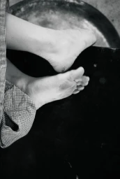 Close-up black and white photograph of bare feet resting over a dark surface beside fine-check patterned trouser fabric.