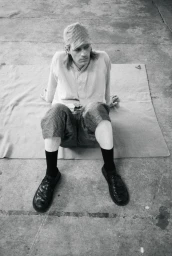 Model wearing a patterned cap, light shirt, patterned shorts, black socks, and shoes seated on a mat on concrete.
