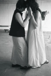 Two women standing face-to-face touching palms and foreheads in monochrome, wearing a sleeveless maxi dress and a dark vest.