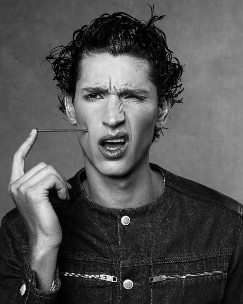 Black and white portrait of a young adult male with curly hair wearing a denim jacket, holding a small metal rod near his face.