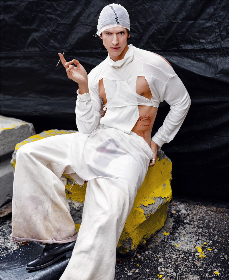 Fashionable adult male sitting on a yellow painted rock, wearing tattered white clothing, holding a cigarette indoors against a dark backdrop.