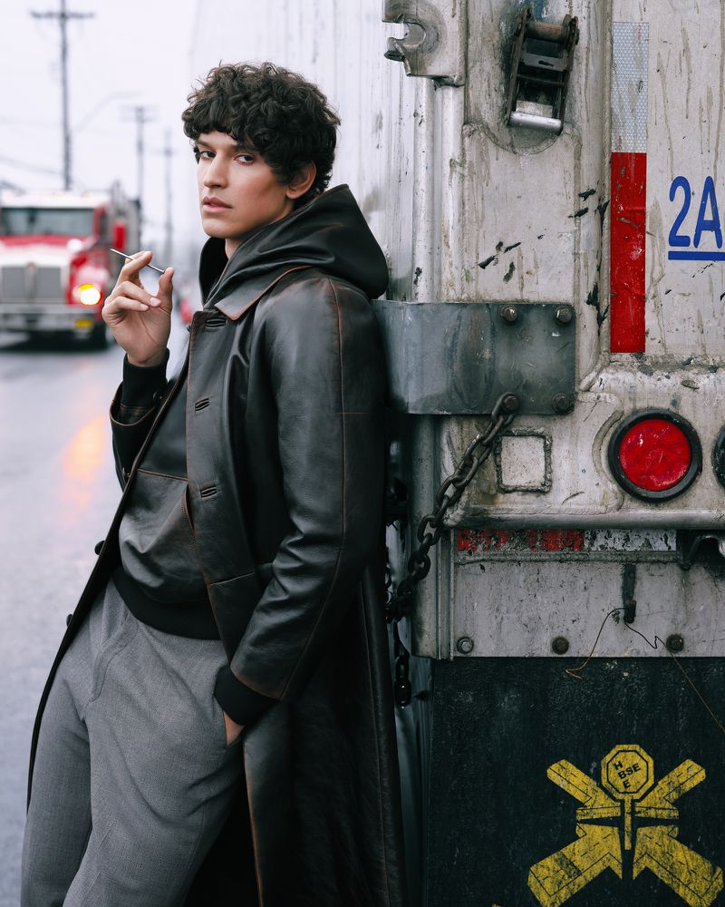 Man in a leather coat leans against a truck on a city street, holding a cigarette. A red truck is visible in the background.