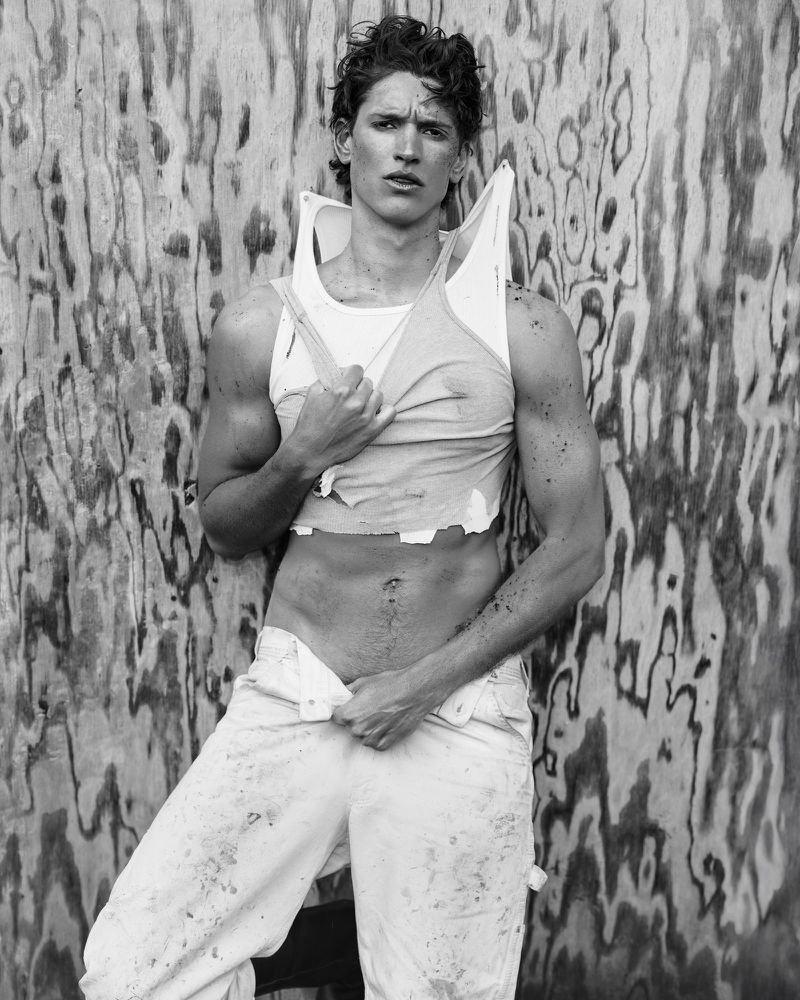 Black and white portrait of a tanned adult man in a torn white tank top and white trousers, posing against a textured wall.