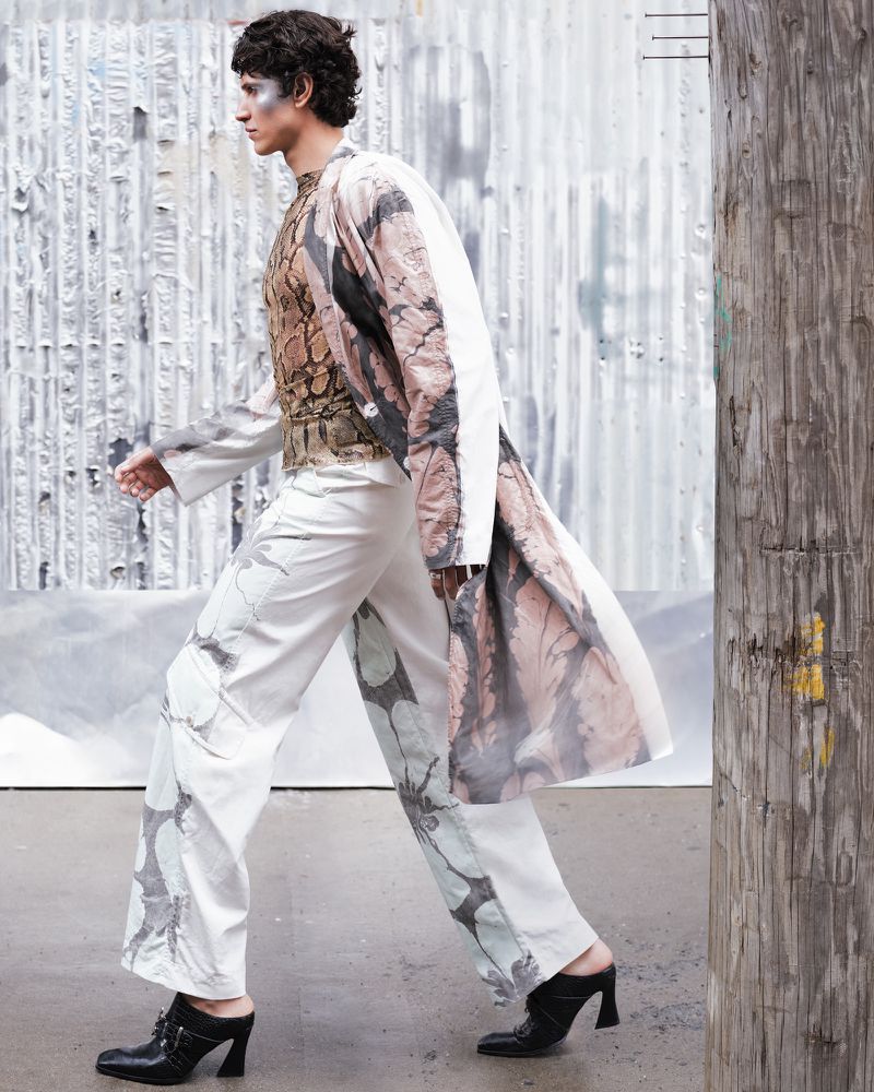 Editorial fashion shot of a slender man walking with white printed pants, reptile patterned top, jacket, and black heeled mules in urban setting.