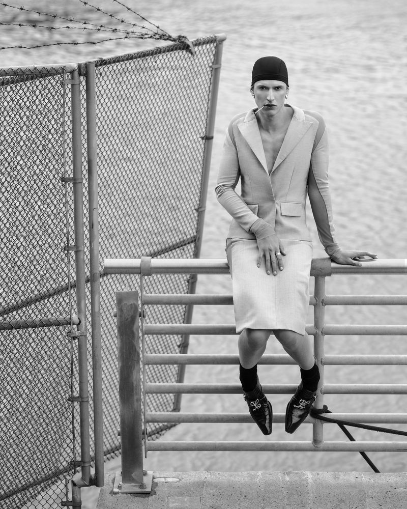 Monochrome portrait of a person with a cap, wearing a blazer and skirt, sitting on a metal railing against a water and chain-link fence background.