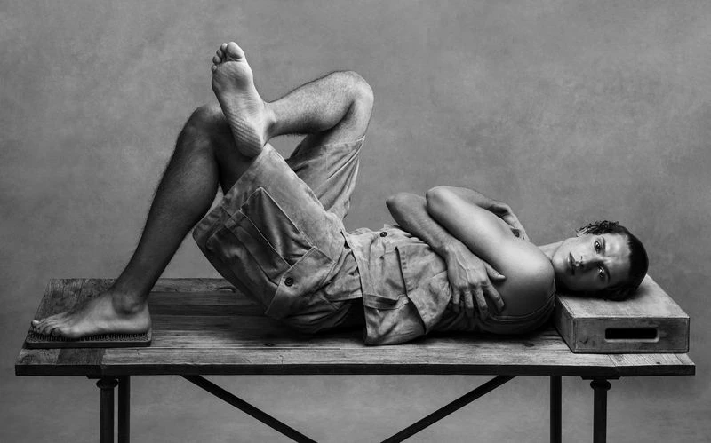 An adult man poses on a wooden table with nail board in a black and white studio shot, wearing shorts and a tank top with arms crossed.