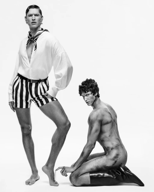 Two male models in a studio setting; one is standing in striped shorts, and the other is kneeling on a dark cushion wearing boots.