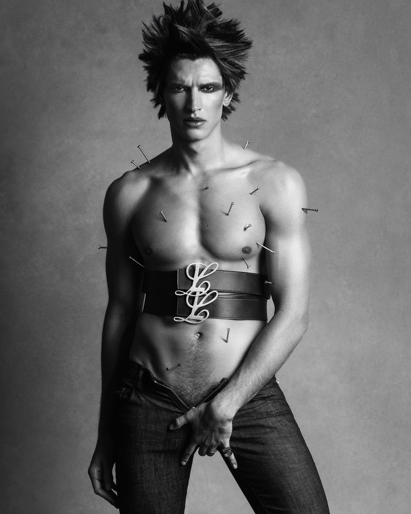 Black and white studio shot of a man with spiky hair, nails on his chest, wearing jeans, and a wide belt with decorative details.