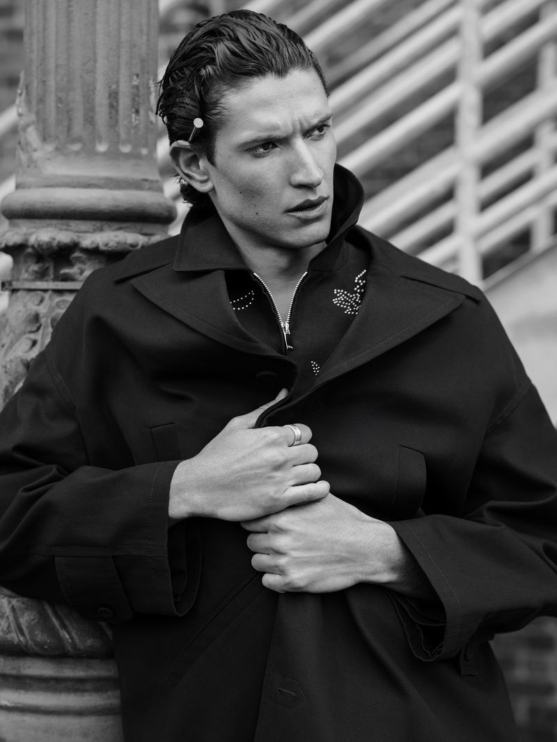 Black and white portrait of a young adult male wearing a dark overcoat, standing outdoors with architectural column background. Fashion editorial.