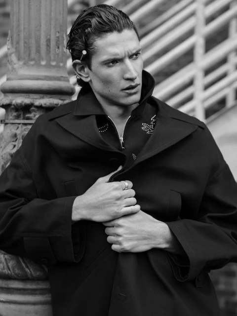 Black and white portrait of a young adult male wearing a dark overcoat, standing outdoors with architectural column background. Fashion editorial.