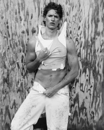 Monochrome fashion portrait of a male model holding a distressed white tank top and unbuttoning trousers against a plywood background.
