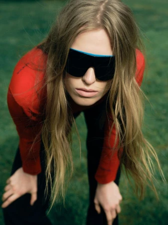 Leanne De Haan wearing oversized black shield sunglasses and a red long-sleeve top bending forward outdoors.