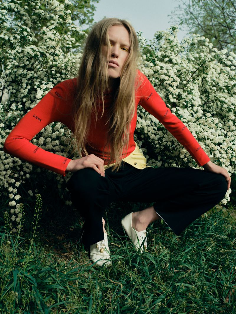 Model Leanne De Haan crouching in grass wearing a bright red long-sleeve top and black trousers before blooming white bushes.