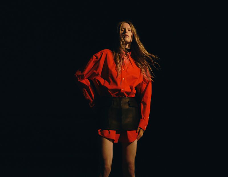Model Leanne De Haan wears an oversized bright red button-down shirt and black leather mini skirt against a dark background.