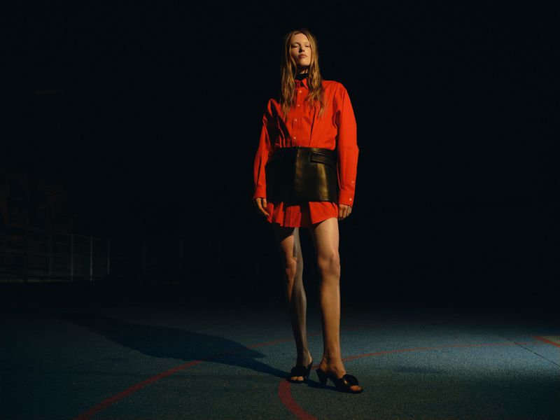 Model Leanne De Haan wearing an oversized red shirt and black leather skirt standing under spotlight on a dark sports court.