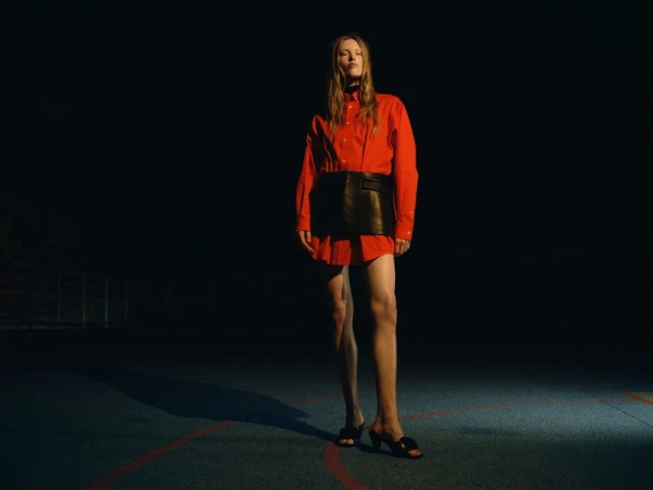Model Leanne De Haan wearing an oversized red shirt and black leather skirt standing under spotlight on a dark sports court.