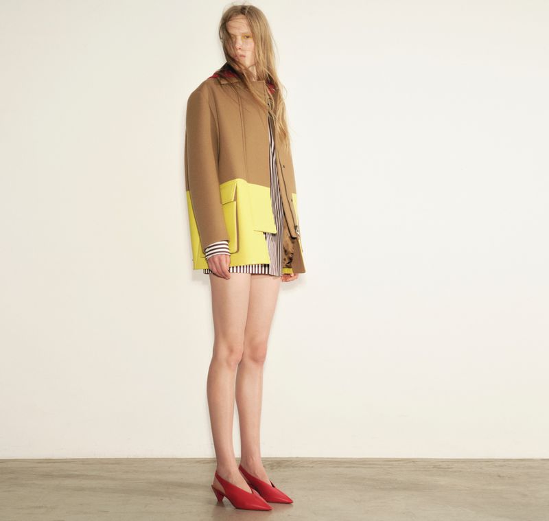 Model Leanne De Haan wearing a camel and yellow color-blocked jacket with red slingback heels against a white studio background.