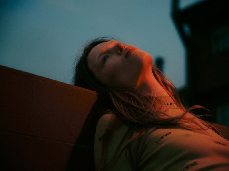 Model Leanne De Haan leaning back against a wall at dusk, wearing a beige patterned high-neck Loewe top illuminated by warm light.