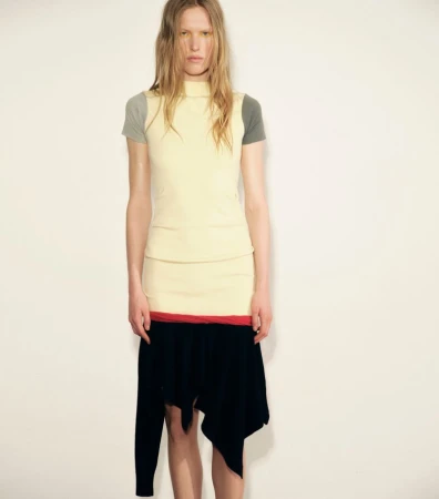 Model Leanne De Haan posing in a layered pale yellow top and black asymmetrical skirt against a plain studio background.