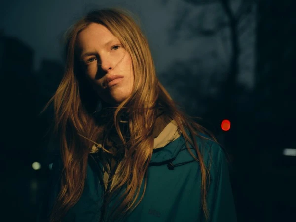 Model Leanne De Haan illuminated by warm directional light wearing a teal jacket against a dark night street background.