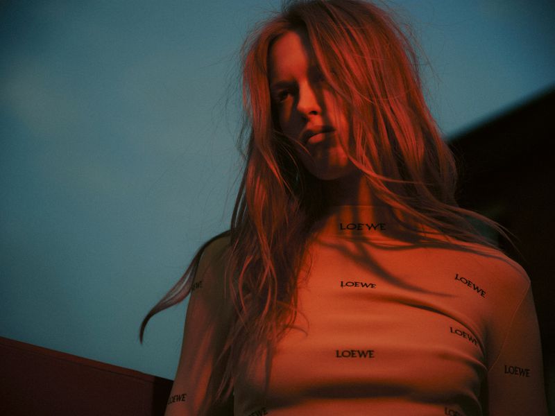 Model Leanne De Haan wearing a printed Loewe top under dramatic warm amber lighting against a deep blue dusk sky.
