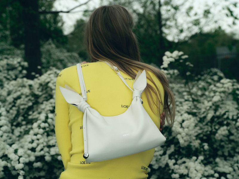 Model seen from behind wearing a bright yellow logo top and a white knotted leather shoulder bag amid blooming bushes.