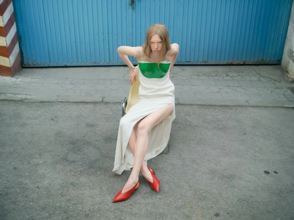 Model Leanne De Haan seated in a white slip dress with a green bodice insert and red pointed shoes before a blue door.