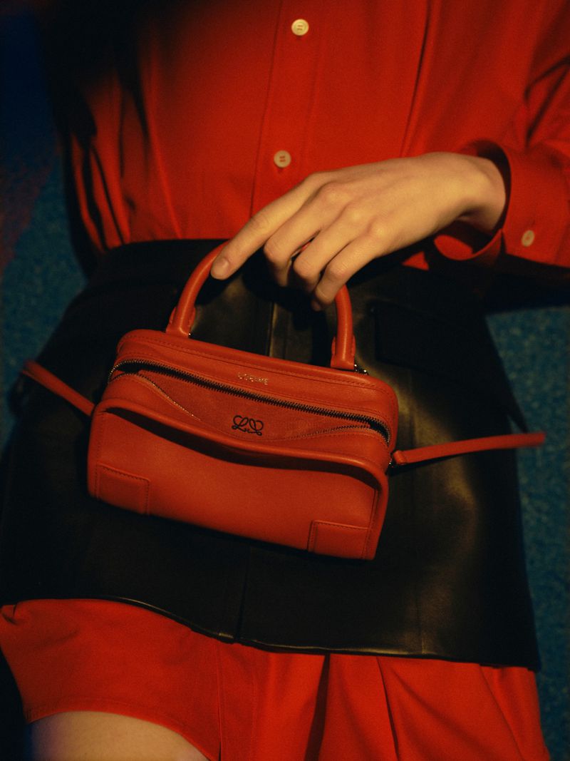 Model wearing a bright red button-up shirt and black leather skirt holding a small red leather handbag with a top handle.