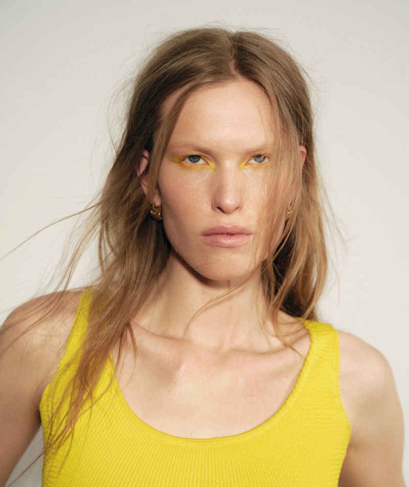 Model Leanne De Haan wearing bold yellow graphic eye makeup and a yellow ribbed tank top against a pale backdrop.