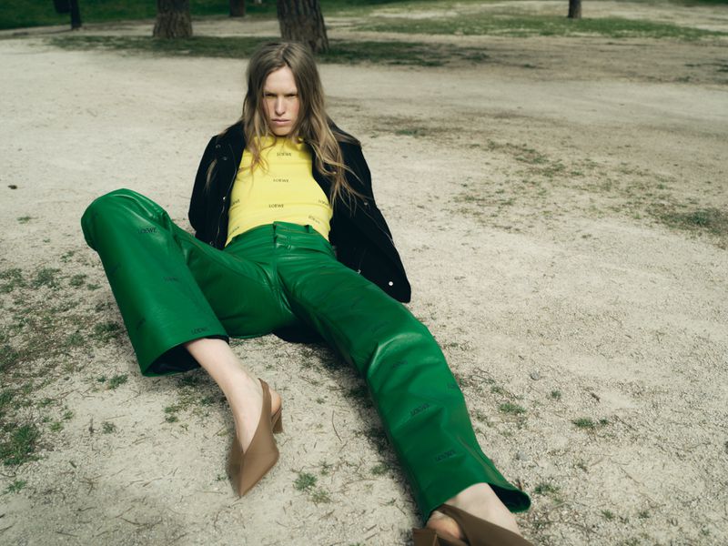Model Leanne De Haan poses reclining outdoors wearing bright green leather trousers, a yellow top, and a black jacket.