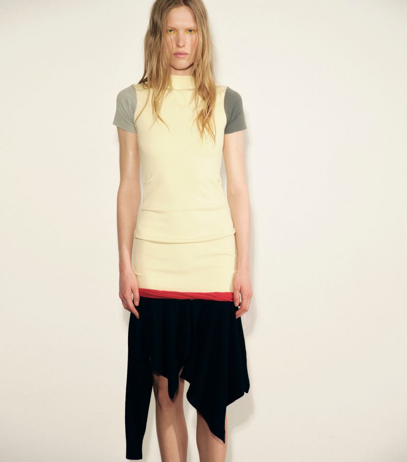 Model Leanne De Haan posing in a layered pale yellow top and black asymmetrical skirt against a plain studio background.