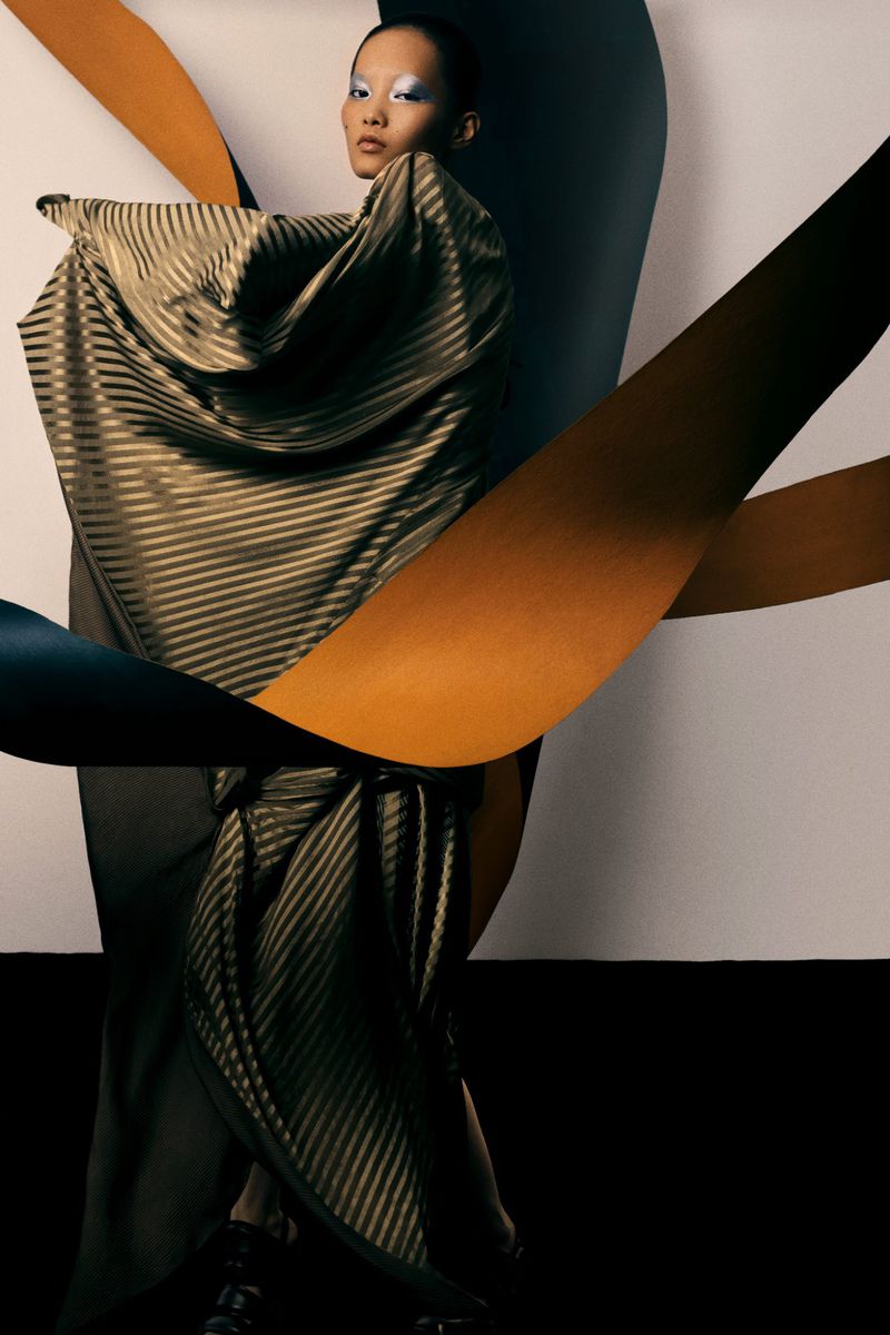 Asian female model in a gold striped gown poses against a gradient paper background, showcasing fashion and studio photography.